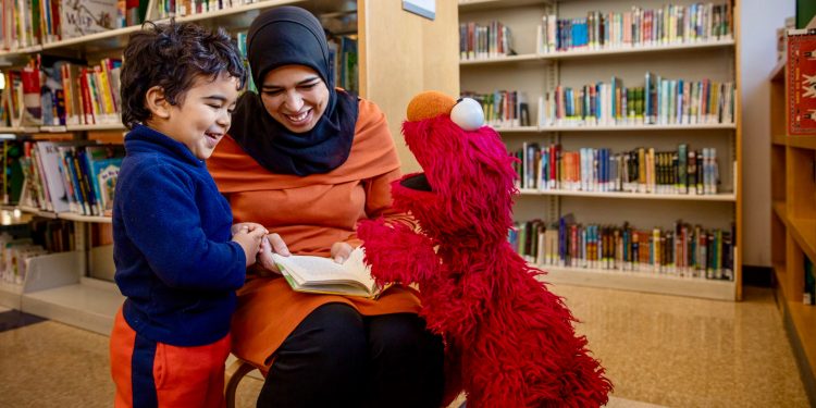 A woman in a children's library reading to Elmo and a child.